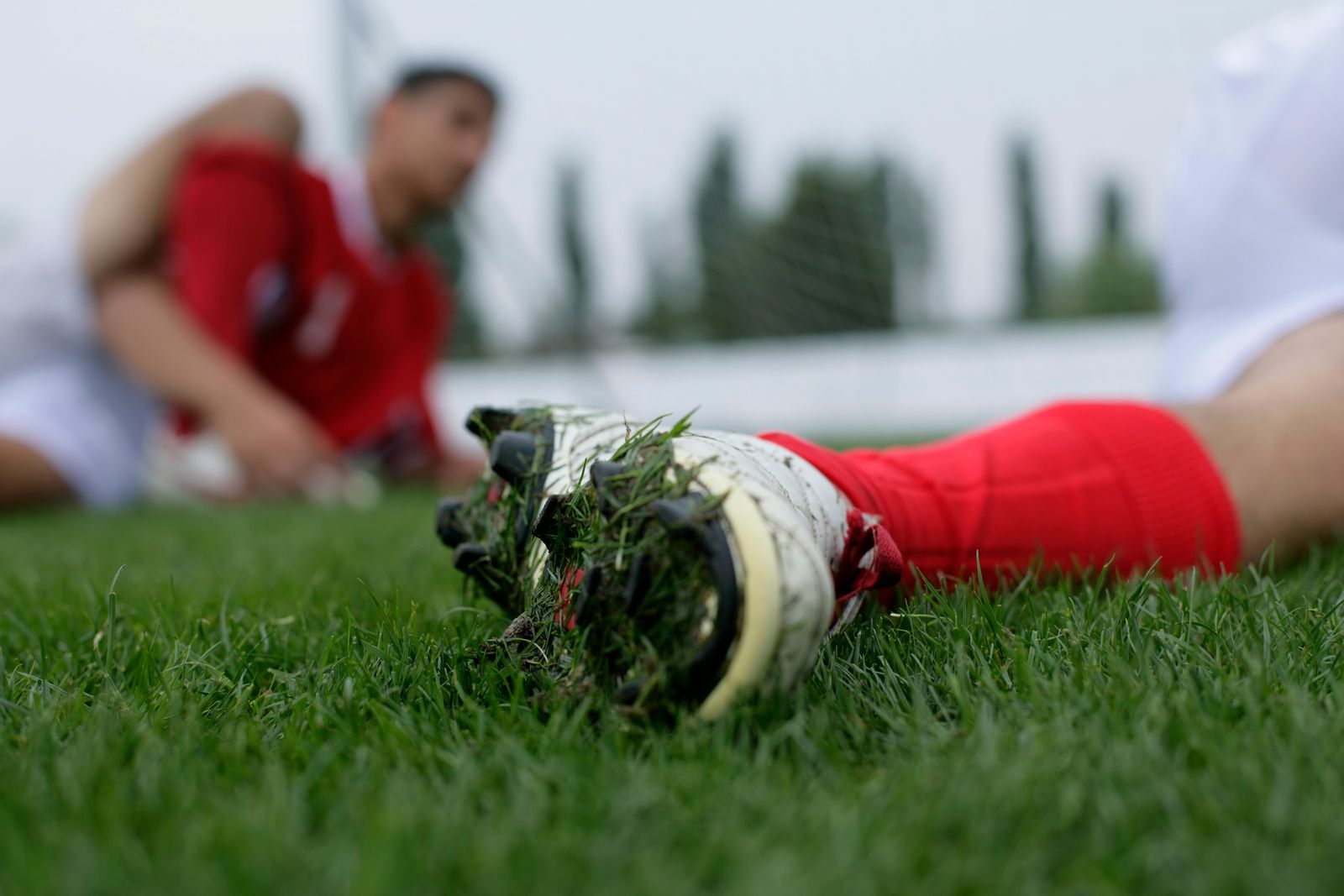 Close-up of a football boot's studs caught in grass, with a red-socked teammate stretching in the blurred background.