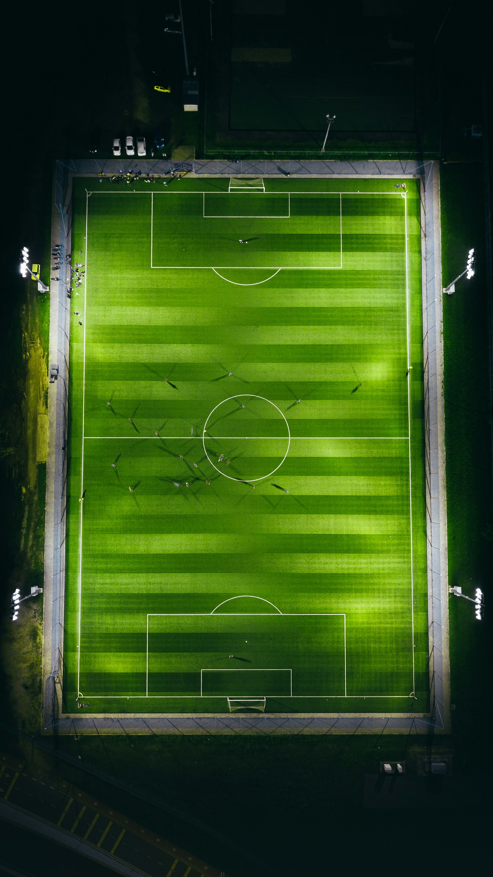 An aerial night view of a floodlit green football pitch mid-match, with players' shadows stretching across the centre circle.