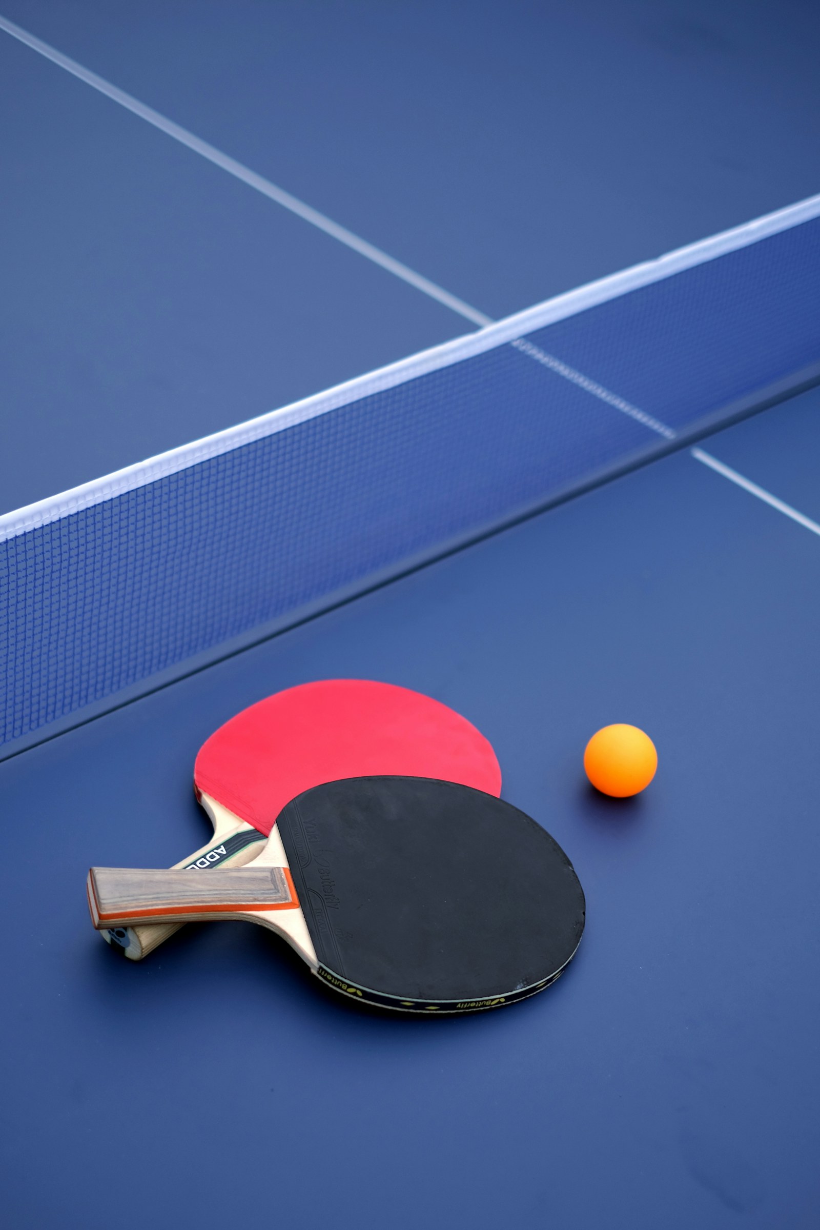 Two table tennis paddles, one red and one black, rest beside an orange ball on a blue table next to the net.